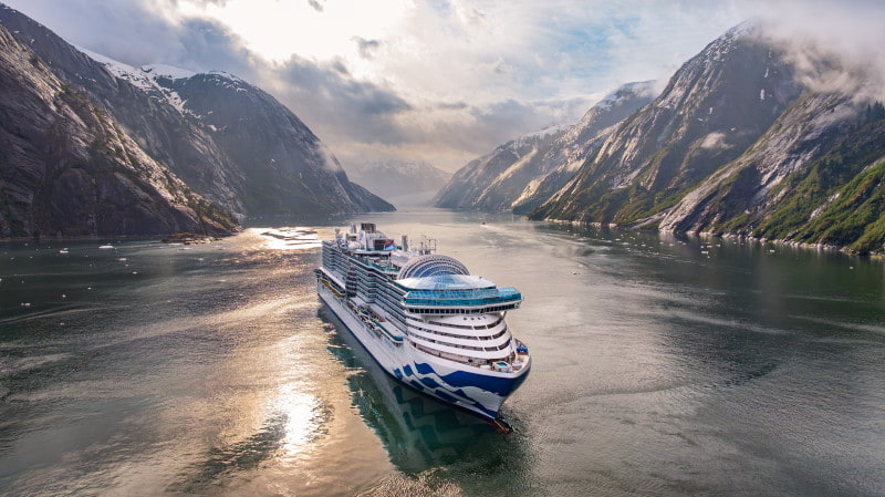 cruise ship navigating a narrow fjord surrounded by steep, mist-covered mountains at sunrise.