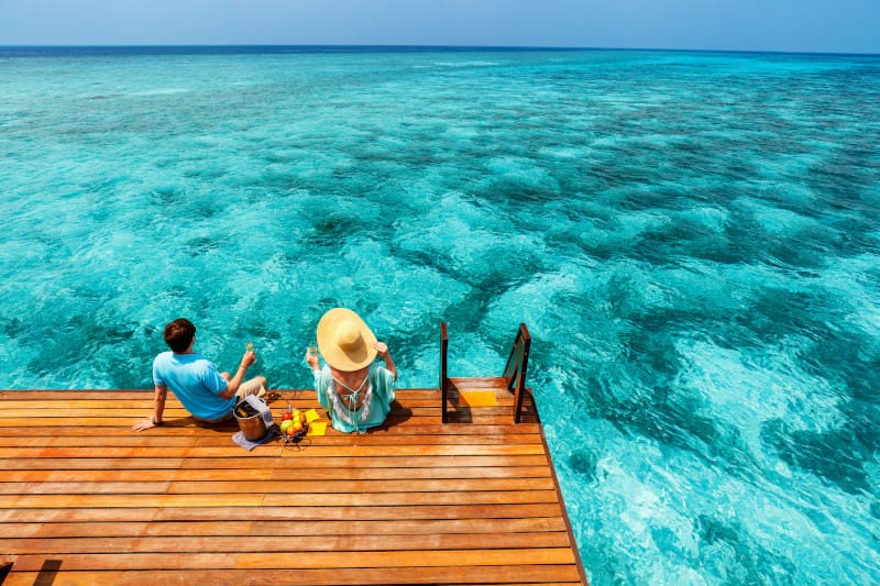 couple relaxing on an overwater wooden deck with drinks, overlooking crystal-clear turquoise water