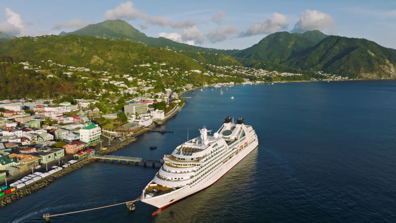 Aerial view of a small cruise ship docked along a lush green Caribbean coastline near a hillside town.