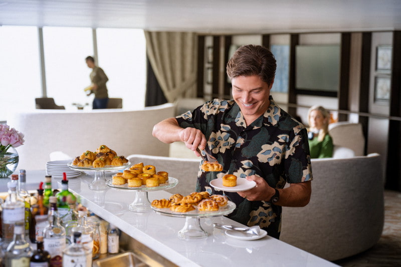 Guest browsing a tray of pastries at an onboard lounge bar, with a shipboard bar and lounge seating in the background.