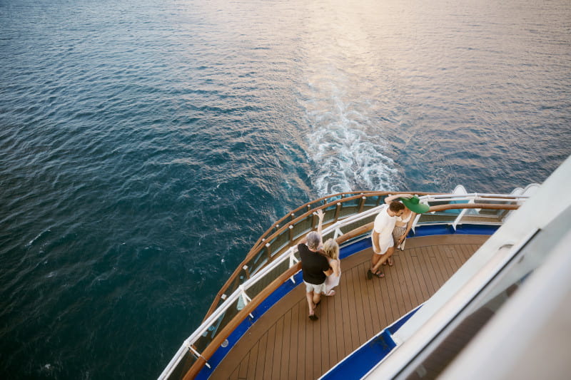 Couple standing at the aft railing of a cruise ship, watching the wake trial across the ocean at golden hour.
